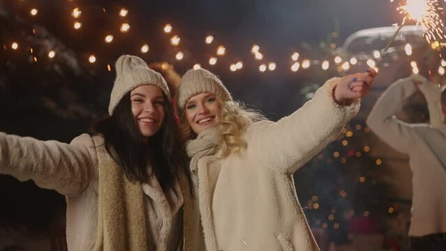 two beautiful women swaying sparklers and smiling to camera on town square happy New year and Xmas