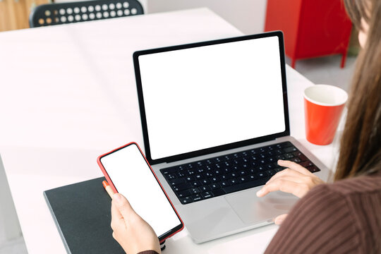 Over the shoulder mockup image of young adult businesswoman working on laptop computer and mobile phone with white blank screen for advertising text