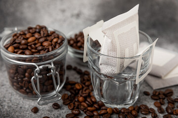 Drip coffee bags, coffee beans and a glass cup on a texture table. Ground coffee for brewing in a cup, paper bag drip coffee filter packaging. Place to copy. Place for text.