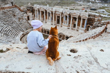 A little girl with her Shiba Inu dog sits on top of the amphitheater and looks into the distance....