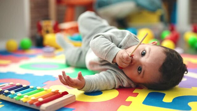 Adorable hispanic baby holding xylophone stick lying on floor at kindergarten