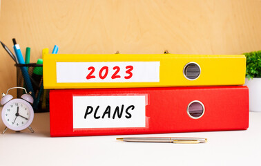 Red and yellow folders lie on the office table next to the clock and pen. Inscriptions on 2023 and PLAN folders.