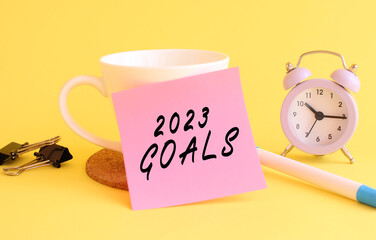 Pink paper with the text 2023 GOALS on a white cup. Clock, pen on a yellow background.