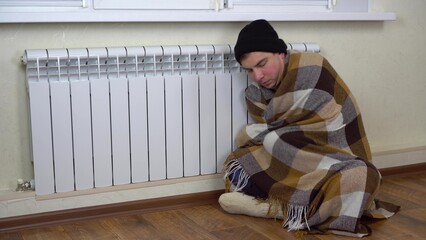A young man is sitting on the floor by the radiator covered with a blanket. A man in a hat and...