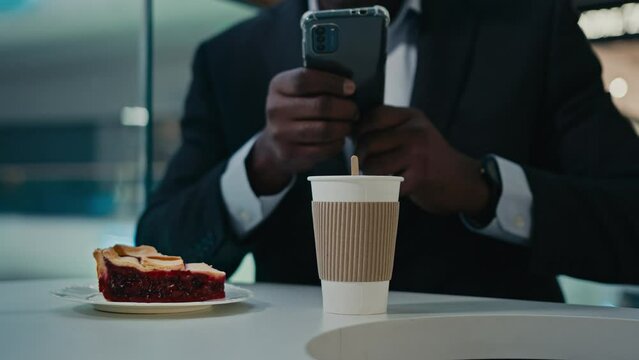 Unrecognizable man African businessman entrepreneur with mobile phone browsing chatting make picture photo blogger vlogger male at table with smartphone in cafe with cake coffee messaging in cafeteria