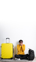 Young tourist traveler with a yellow suitcase covering his mouth with his hands keeps a secret while sitting on a white background. Air flight journey concept