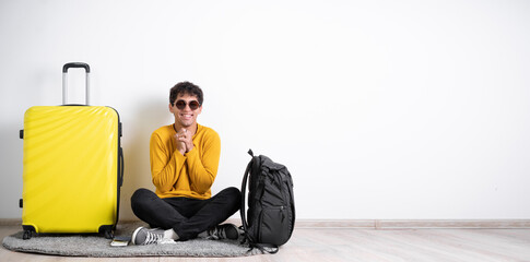 Young guy traveler with a suitcase satisfied with a positive expression sits isolated on a white background holds his hands near his chest