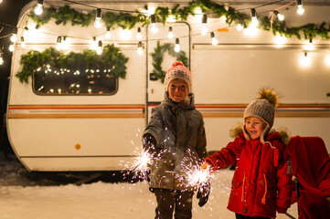 Caucasian red-haired boys hold sparklers by the trailer. Two brothers are celebrating Christmas on a trip.