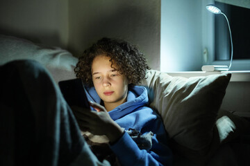 Teenage girl uses a smartphone in a dark room without electricity and heating sits under blanket with led light from power bank. Closeup portrait.