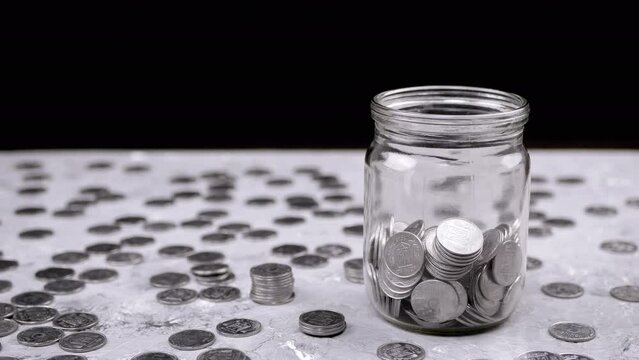 Hand Throws One Five-Kopeck Ukrainian Coin into a Glass Jar Filled with Coins. A Pile of scattered coins is on table. Stock. Piggy bank. Black background. Financial crisis, budget savings. Poverty.
