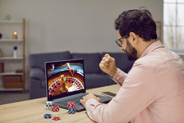 Young man gambling in virtual online casino on Internet. Happy guy sitting at desk table with poker...