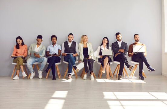 Group of different multiethnic people sitting in row in waiting room before interview or meeting. People read resumes, view documents and use digital devices while sitting in empty lobby with sunlight