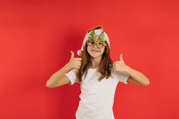 Close-up portrait of a teenage girl in a santa cap and Christmas glasses on a red background, shows fingers up