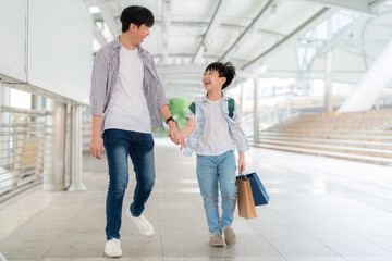 Young Asia boy holding colorful shopping bag and walking with father in the shopping central area. Enjoyment on shopping and happy with gift during end of the year. Mid year sale and holiday concept