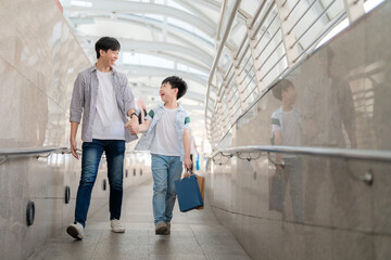 Young Asia boy holding colorful shopping bag and walking with father in the shopping central area. Enjoyment on shopping and happy with gift during end of the year. Mid year sale and holiday concept