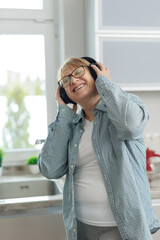 An adult relaxing woman in wireless headphones listens to music and dances in the kitchen. A woman in a blue shirt smiles and dances.