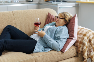 Obraz premium Elderly woman 50s years old wears casual clothes lies on sofa use laptop computer watch movie film comedy drink wine laugh stay at home flat rest relax spend free spare time in living room indoor