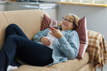 Obraz premium Elderly woman 50s years old wears casual clothes lies on sofa use laptop computer watch movie film comedy drink wine laugh stay at home flat rest relax spend free spare time in living room indoor