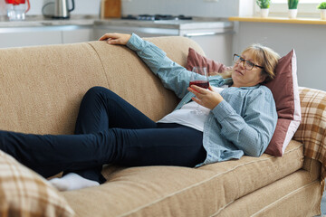Obraz premium Elderly woman 50s years old wears casual clothes lies on sofa use laptop computer watch movie film comedy drink wine laugh stay at home flat rest relax spend free spare time in living room indoor