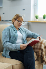 Portrait of mature woman in eyeglasses reading book on sofa at home