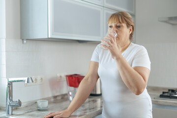 Mature woman in white t-shirt and gray leggings drinks clean water while standing in the kitchen.