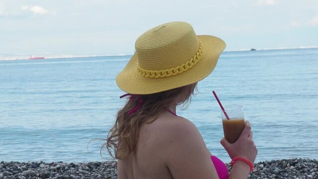 Pretty woman drinks refreshing cocktail yellow straw hat and pink bathing suit. Female enjoys a drink near sea on the beach with blue water. Girl chilling with beverage in tropical sun. Vacation conce