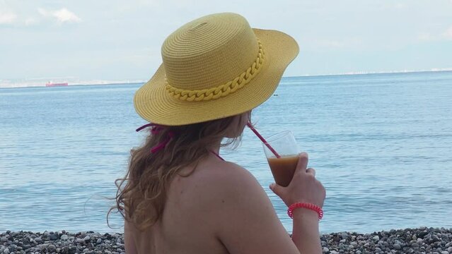 Pretty woman drinks refreshing cocktail yellow straw hat and pink bathing suit. Female enjoys a drink near sea on the beach with blue water. Girl chilling with beverage in tropical sun. Vacation conce