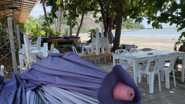 Beachfront view folded purple umbrella, plastic chairs, tree near sandy beach on sunny day. Hua Hin beach
