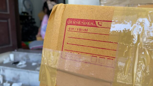 Close up cardboard box package with sender and postcode word screen on parcel with blurred person working in background