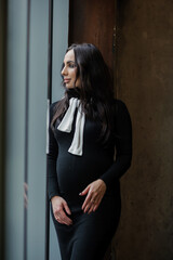 A pregnant woman in a black dress stands near the window