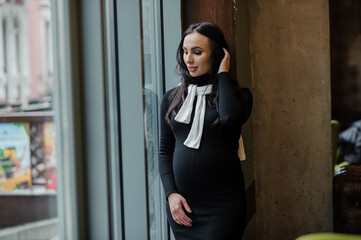 A pregnant woman in a black dress stands near the window