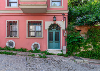 Fototapeta na wymiar Exterior facade of old residential building with walls painted in pink, wrought iron windows, and green decorated door, at a narrow cobblestone street, with trees, Balat district, Istanbul, Turkey