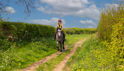 Young woman and her grey horse canter along a bridlepath  on a sunny day , enjoying the freedom to move at speed and enjoy nature.