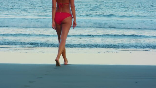 Female in a red bekini goes for a swim. Vacation travel bikini woman walking on beach into water. Lady with slim sexy body on sand beach in going swimming in perfect ocean. Luxury destination.