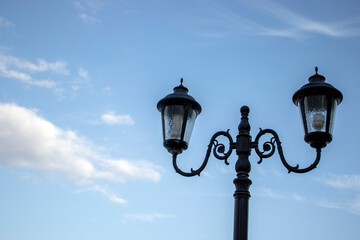 Fototapeta na wymiar Openwork street lamps against the blue sky during the day. The outlines of ornate lighting lanterns, side view. Figured lanterns of street lighting with two plafonds.