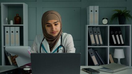Young doctor arabian muslim religion woman doing paperwork at clinic make scientific research read paper documents medical insurance female physician medic use laptop modern technology in healthcare