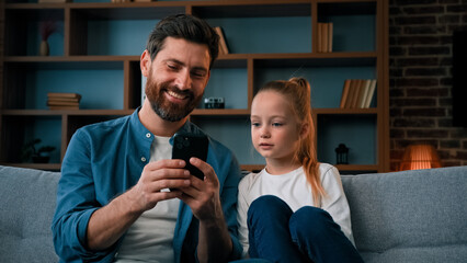 Cute small baby girl child resting on cozy couch with caring young father man playing game on smartphone watching media content on phone using mobile apps shopping online taking family selfie together