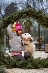 Portrait of a cute woman with her dog hugs together in a big Christmas wreath at backyard. Concept of love and coziness during winter holidays