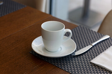 Empty Glass white tea cup standing on desk background, close up. Coffee porcelain mug with saucer on wooden table in the morning sun light. Breakfast beverage. Simple white tableware, selective focus