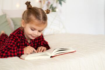 cute little girl in red Christmas pajama lying on bed reading book