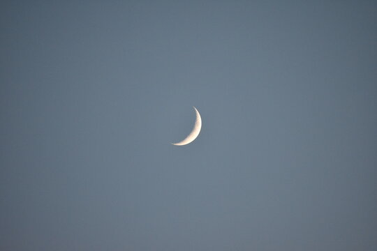 Waxing Cresent moon early on a clear evening backdropped against a light blue sky. Craters visible on surface. 