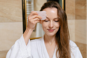A young woman with wet hair in a white bathrobe is massaging her face with a quartz gouache jade...
