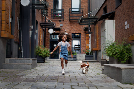 Happy Black Child Girl With Dog Running On City Street Outside. African American Little Model With Cocker Spaniel