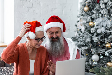 Happy loving senior couple having fun on New Year party near Christmas tree. Mature man and woman smiling, making video call. Concept of active life and leisure of pensioner