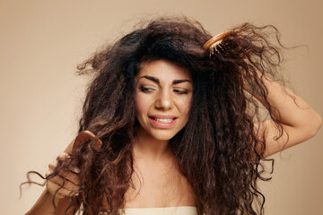 DAMAGED HAIR. HAIR PROBLEMS CONCEPT. Irritated suffering awesome curly Latin lady with hairbrush pulling combing hair posing isolated on pastel beige background, look aside. Copy space, good offer