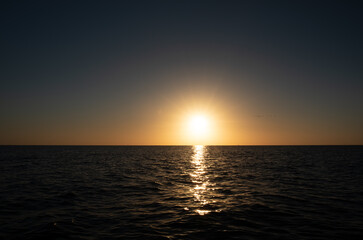 The sun sets over the horizon. In the foreground the sea reflecting the sun. A flock of birds flies towards the sun. The sky and the water are dark.