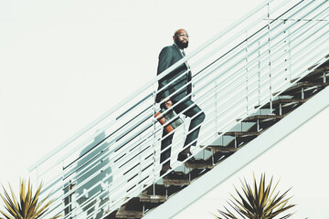A bald black bearded man walks up the stairs. The African male is ascending a white metallic staircase on a shiny day, wears a dark suit and holds a leather purse in a very clean and cactus aesthetic - Powered by Adobe
