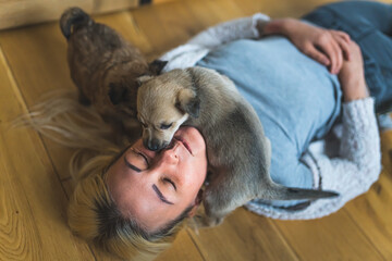 Young adult caucasian woman lying down on wooden floor with closed eyes with a brown mixed-breed puppy dog on her neck and other puppy dog standing on her blond hair. High quality photo