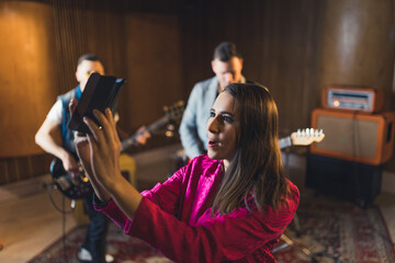 cheerful singer-woman streaming a live video during repetition, other group members in the background music studio. High quality photo