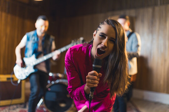 dark-haired enthusiastic woman singing into a microphone and posing for a camera her band playing in the background. High quality photo